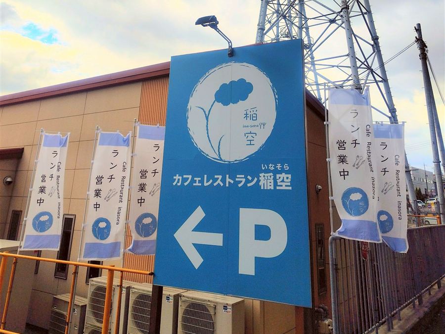 青い看板。稲空のロゴと「カフェレストラン稲空」の文字、駐車場を示す矢印があり、まわりに「ランチ営業中」ののぼり旗が立っています。